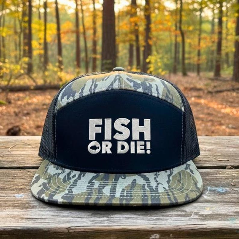 Puede incluir: Gorra de camionero con estampado de camuflaje, con la parte trasera de malla negra y un panel frontal azul marino. El panel presenta el texto blanco "FISH OR DIE!" con un gr&aacute;fico de pez. La gorra est&aacute; sobre una superficie de madera.