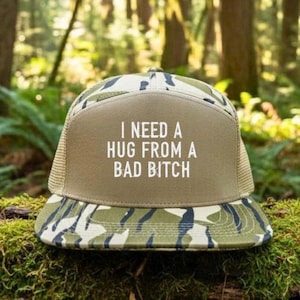 May include: A trucker hat with a tan front panel and a camouflage brim. The hat features the text "I NEED A HUG FROM A BAD BITCH" in white block letters. The hat has a mesh back and a curved brim.