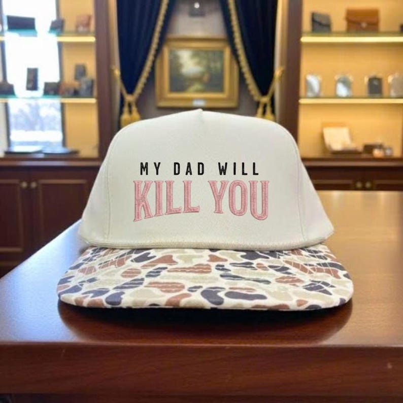 May include: A white baseball cap with the text "MY DAD WILL KILL YOU" in black and pink. The brim features a camouflage pattern in shades of brown, tan, and gray. The cap is sitting on a dark brown surface.
