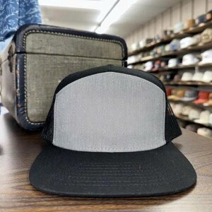 May include: A black trucker hat with a grey front panel and a black peak. The hat has a mesh back and is on a wooden surface. A vintage-style suitcase is in the background.