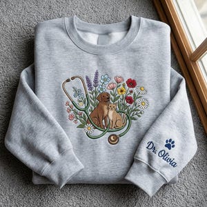 May include: A light gray sweatshirt featuring a colorful illustration of a dog and cat with a stethoscope and flowers. The sleeve has the text "Dr. Olivia" and a paw print.