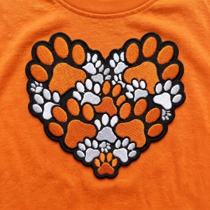 Może przedstawiać: Pomarańczowy t-shirt z dużą, haftowaną naszywką w kształcie serca. Wzór składa się z licznych, nakładających się na siebie pomarańczowych i białych odcisków łap, obszytych czarną nicią, tworząc gęsty i teksturowany motyw serca.