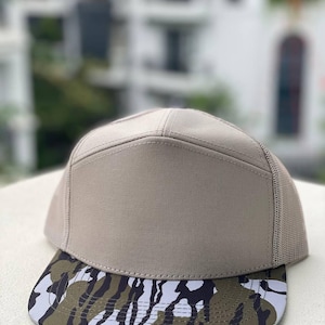 May include: A tan baseball cap with a camouflage brim. The cap has a mesh back and a structured front. The brim features a pattern of black, white, and olive green shapes. The cap is sitting on a white surface.