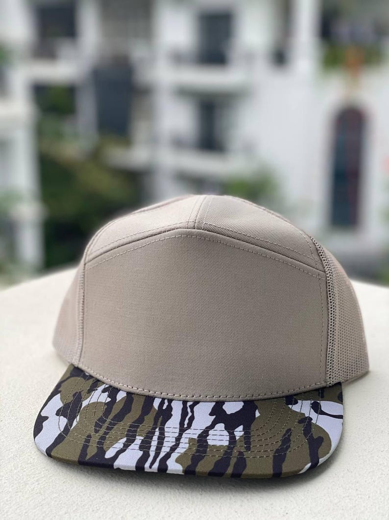 May include: A tan baseball cap with a camouflage brim. The cap has a mesh back and a structured front. The brim features a pattern of black, white, and olive green shapes. The cap is sitting on a white surface.