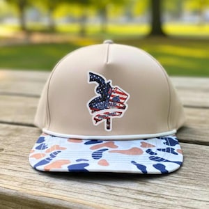 Peut inclure: Casquette de baseball beige avec une visière camouflage blanche, bleue et pêche. Le chapeau présente un écusson avec un motif patriotique, comprenant une silhouette de pistolet, un drapeau américain et le texte "Stand Your Ground".