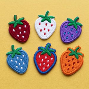 Op de afbeelding: Zes geborduurde aardbeienpatches in verschillende kleuren: rood, wit, paars, blauw, rood met blauwe rand en oranje. Elke patch heeft een groene steel en witte zaaddetails, perfect voor knutselen of het versieren van kleding.