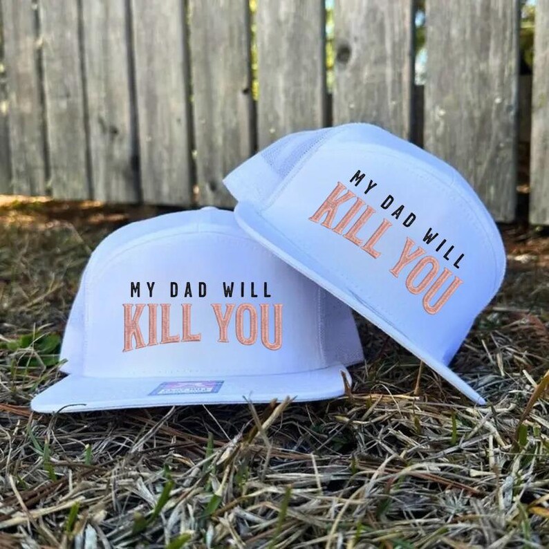 May include: Two white trucker hats with the text "MY DAD WILL KILL YOU" embroidered in orange. The hats have a white mesh back and a flat brim. The hats are resting on a bed of dry grass.