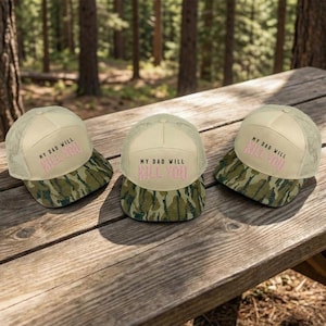 May include: Three beige trucker hats with a camouflage brim are displayed on a wooden picnic table. The hats have the text "MY DAD WILL KILL YOU" in pink. The background is a blurred forest scene.