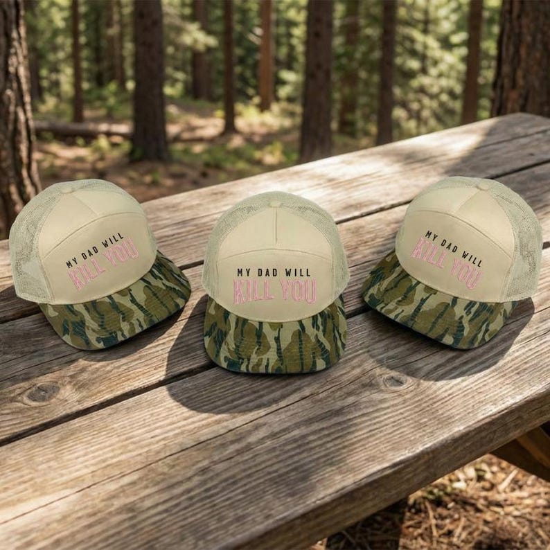 May include: Three beige trucker hats with a camouflage brim are displayed on a wooden picnic table. The hats have the text "MY DAD WILL KILL YOU" in pink. The background is a blurred forest scene.