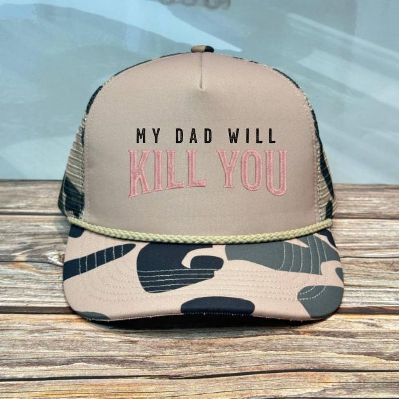 May include: A trucker hat with a tan front panel and a mesh back. The hat features the text "MY DAD WILL KILL YOU" in black and pink embroidery. The brim has a camouflage pattern.