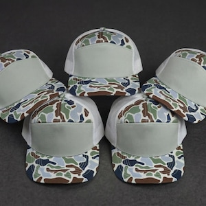 May include: A collection of five trucker hats with a camouflage pattern on the front and a solid gray front panel. The hats have a white mesh back and a curved brim. The hats are arranged in a circular pattern on a dark gray surface.