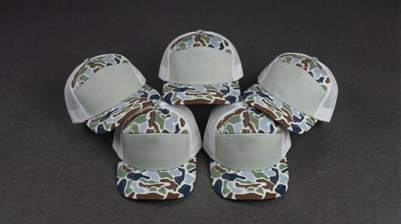 May include: A collection of five trucker hats with a camouflage pattern on the front and a solid gray front panel. The hats have a white mesh back and a curved brim. The hats are arranged in a circular pattern on a dark gray surface.