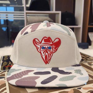 May include: A beige trucker hat with a camouflage pattern on the bill and crown. The hat features a red cowboy graphic with a blue bandana and eyes. The hat has a white mesh back.