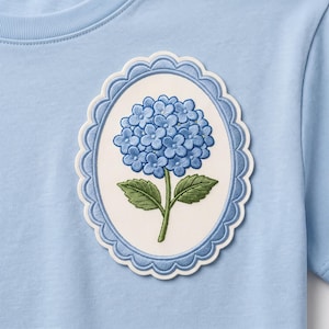 May include: A light blue t-shirt featuring an oval-shaped embroidered patch. The patch has a scalloped blue border and a white center displaying a blue hydrangea flower with green leaves. The embroidery is detailed with visible stitching.