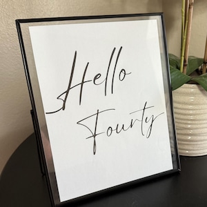 May include: A framed print with the words "Hello Forty" in elegant black script on a white background. The frame is black and sits on a dark surface. A decorative plant is in the background.