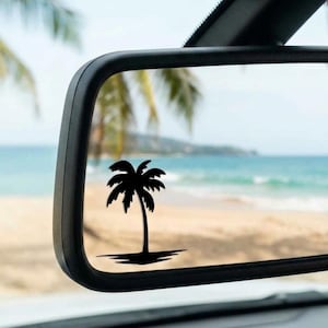 May include: A black palm tree decal applied to the surface of a rectangular vehicle rearview mirror. The decal features a single palm tree silhouette with a small shadow at the base, contrasting against the reflection of a beach and ocean scene.