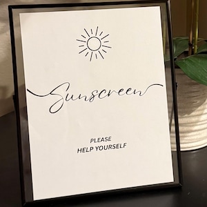 May include: A framed white sign with black text that reads "Sunscreen" in cursive, with a sun illustration above the word. Below the word, the sign reads "PLEASE HELP YOURSELF". The frame is black.