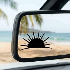 May include: A black vinyl decal of a semi-circular sun with radiating rays applied to the surface of a rectangular car rearview mirror. The design features a solid half-circle base with thin, pointed lines extending outward in a fan shape.