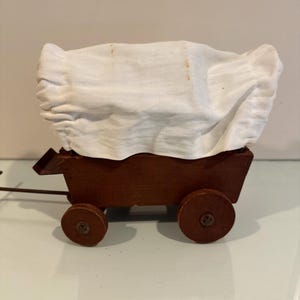 May include: A small wooden covered wagon, featuring a brown body and four circular wheels. It is covered with a white fabric top. This miniature wagon could be a vintage toy or a decorative piece.