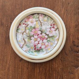 May include: Circular coaster with a floral pattern. It has a cream-coloured border with a gold trim. The centre of the coaster displays pink flowers, green leaves, and red berries on a light grey background.