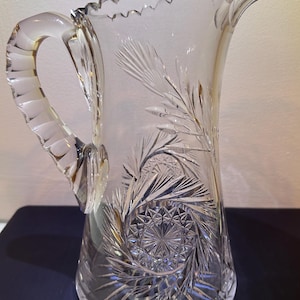 Könnte beinhalten: Klarer Glaskrug mit dekorativem Griff und gewelltem Rand. Der Krug weist aufwendige geätzte Muster auf, darunter ein Spiraldesign am Boden und farnartige Motive. Ein klassisches Stück zum Servieren von Getränken.