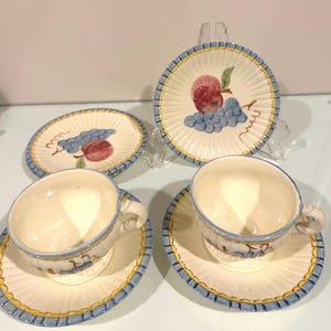 Puede incluir: Un juego de cuatro tazas y platillos de cerámica. Las tazas y platillos de color blanquecino tienen un borde azul y amarillo. Cada platillo presenta un diseño pintado de una manzana roja y uvas azules. Las tazas tienen un diseño de fruta a juego.