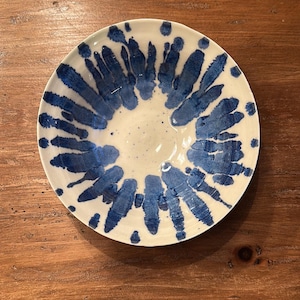 May include: A ceramic bowl with a white interior and a blue, abstract design radiating from the center. The bowl's rim features a similar pattern. The bowl is set on a wooden surface.