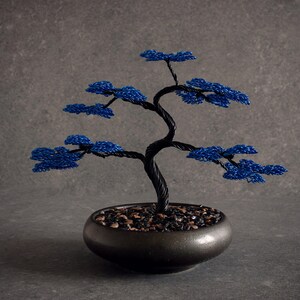 May include: A decorative wire bonsai tree sculpture featuring a twisted black metal trunk and branches with vibrant blue wire foliage. The sculpture is anchored in a round, dark-colored ceramic pot filled with small, dark, polished stones.