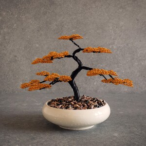 Może przedstawiać: Rzeźba drzewka bonsai wykonana z drutu, z poskręcanym czarnym metalowym pniem i gałęziami z teksturowanym pomarańczowym listowiem. Drzewko jest osadzone w płytkiej, gładkiej białej ceramicznej misie wypełnionej drobnymi, ciemnymi kamykami dekoracyjnymi.