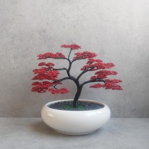 Puede incluir: Un bonsái de alambre hecho a mano con hojas rojas vibrantes y un tronco oscuro. El árbol está en una maceta de cerámica blanca llena de guijarros negros y musgo verde. Un artículo decorativo.