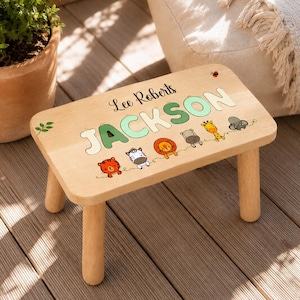 May include: A rectangular light-coloured wooden step stool with four legs. The top surface features the name Lee Roberts Jackson in black and green lettering, accompanied by colourful cartoon illustrations of a tiger, zebra, lion, hippo, giraffe, and elephant.