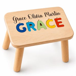 May include: A light-colored wooden stool featuring four cylindrical legs and a rectangular top. The surface displays the name Grace Olivia Martin in black script, with the name GRACE in large, colorful, cutout block letters below it.