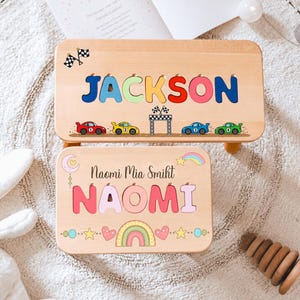May include: Two rectangular wooden step stools with light wood grain. The larger stool features the name JACKSON in colourful letters, race cars, and chequered flags. The smaller stool displays the name NAOMI MIA SMIKT, a rainbow, and hearts.