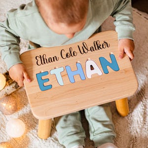 May include: A light-colored wooden step stool featuring a personalized name puzzle on the top surface. The name Ethan Cole Walker is printed in black script, with the name ETHAN displayed in large, blue and white cutout puzzle letters.