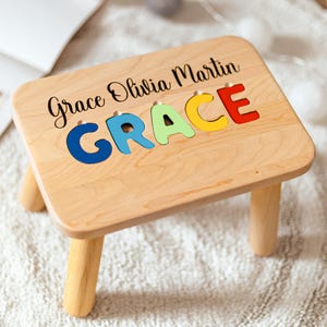 May include: A light-coloured wooden stool with four cylindrical legs. The top surface features the black printed text Grace Olivia Martin above the name GRACE, which is spelled out in large, colourful, individual wooden puzzle-style letters.