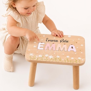May include: A light-colored wooden stool featuring a personalized name puzzle with the letters E-M-M-A in pastel pink and purple. The top surface is decorated with white daisy illustrations and the printed name Emma Rose in black script lettering.