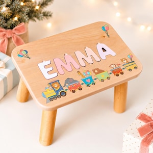 Könnte beinhalten: Ein heller Holzhocker mit dem Namen EMMA in großen, hellrosa ausgeschnittenen Buchstaben. Unter dem Namen befindet sich eine bunte Illustration eines Zuges mit tierischen Passagieren, darunter ein Löwe, ein Elefant, eine Giraffe, ein Bär und ein Affe.