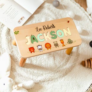 May include: A rectangular wooden step stool featuring the name Lee Roberts and the name JACKSON in large, pastel-colored cutout letters. The surface is decorated with small, colorful illustrations of a tiger, zebra, lion, hippo, giraffe, and elephant.