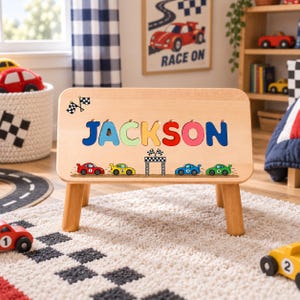 May include: A light-colored wooden puzzle board featuring the name JACKSON in colorful, cutout block letters. Below the name, a graphic shows four small race cars in red, yellow, blue, and green driving toward a checkered finish line.