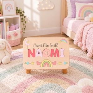 Pode incluir: Um banco de madeira clara tipo puzzle com o nome NAOMI em letras grandes e coloridas. A superfície é decorada com uma lua crescente, um arco-íris, estrelas e corações. O texto Naomi Mia Smiht está impresso acima do nome.