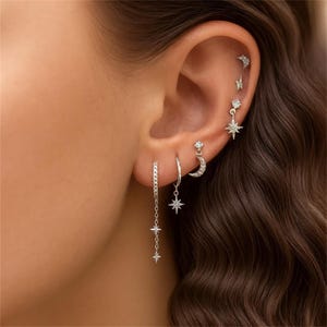 May include: A collection of silver earrings featuring celestial designs. The earrings include star and crescent moon shapes, with some dangling from chains. Several earrings are studded with small, sparkling stones. The earrings are displayed on an ear.
