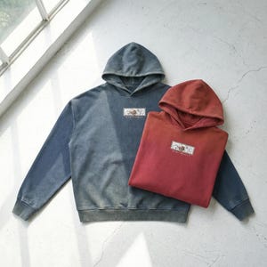 Puede incluir: Dos sudaderas con capucha, una en azul descolorido y la otra en un degradado rojo-naranja. Cada una tiene un pequeño diseño rectangular bordado en el pecho. Las sudaderas están sobre una superficie gris claro cerca de una ventana.