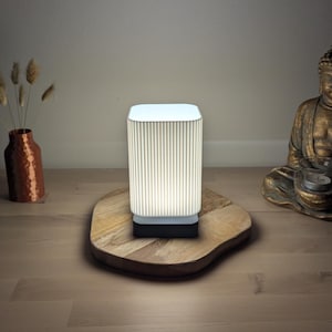 Op de afbeelding: Een moderne, rechthoekige tafellamp met een geribbelde, crèmekleurige kap en een zwarte voet. De lamp staat op een houten, organisch gevormd dienblad. Decoratieve vazen en een bronzen Boeddha-beeld staan op de achtergrond.