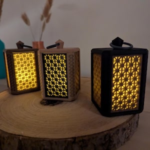 Puede incluir: Tres faroles decorativos con intrincados cortes geométricos, que emiten un cálido resplandor amarillo. Los faroles están hechos de madera y materiales negros, y están colocados sobre una rodaja de madera. Cada farol tiene un asa.