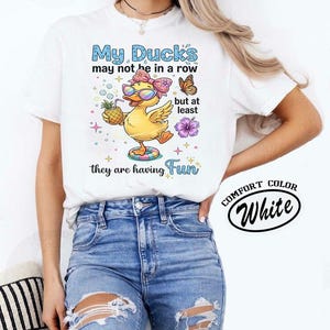 May include: White t-shirt featuring a cartoon duck in sunglasses and a bow, holding a pineapple, and floating on a tube. The text says "My Ducks may not be in a row but at least they are having Fun."