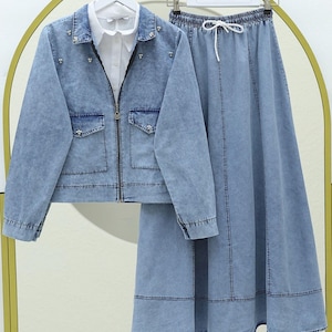 Peut inclure: Ensemble veste et jupe en jean bleu clair. La veste a une fermeture éclair, deux poches avant et des ornements décoratifs. La jupe est longue, de style trapèze, avec une taille élastique. Une chemise blanche à col est visible sous la veste.