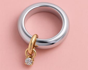 May include: A silver ring with a gold charm and a small, round, clear gemstone. The ring is thick and smooth, with a polished finish. The charm is attached to the ring with a small gold loop. The background is a solid, light pink color.