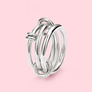 May include: A silver ring with a distinctive design. The ring has multiple bands interwoven with a knot-like feature. The ring is made of a shiny, reflective metal, set against a soft pink background.