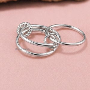 May include: A silver ring with an interlocking design. The ring features three connected bands, with one band having a twisted rope texture. The ring is displayed on a pink surface. The text "Divine Jewels" is visible.