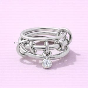 May include: A silver ring with a unique design. The ring features multiple interconnected bands with knot-like details. A small, round, clear gemstone hangs from the center. The words "Divine Jewels" are visible on the left side of the ring.